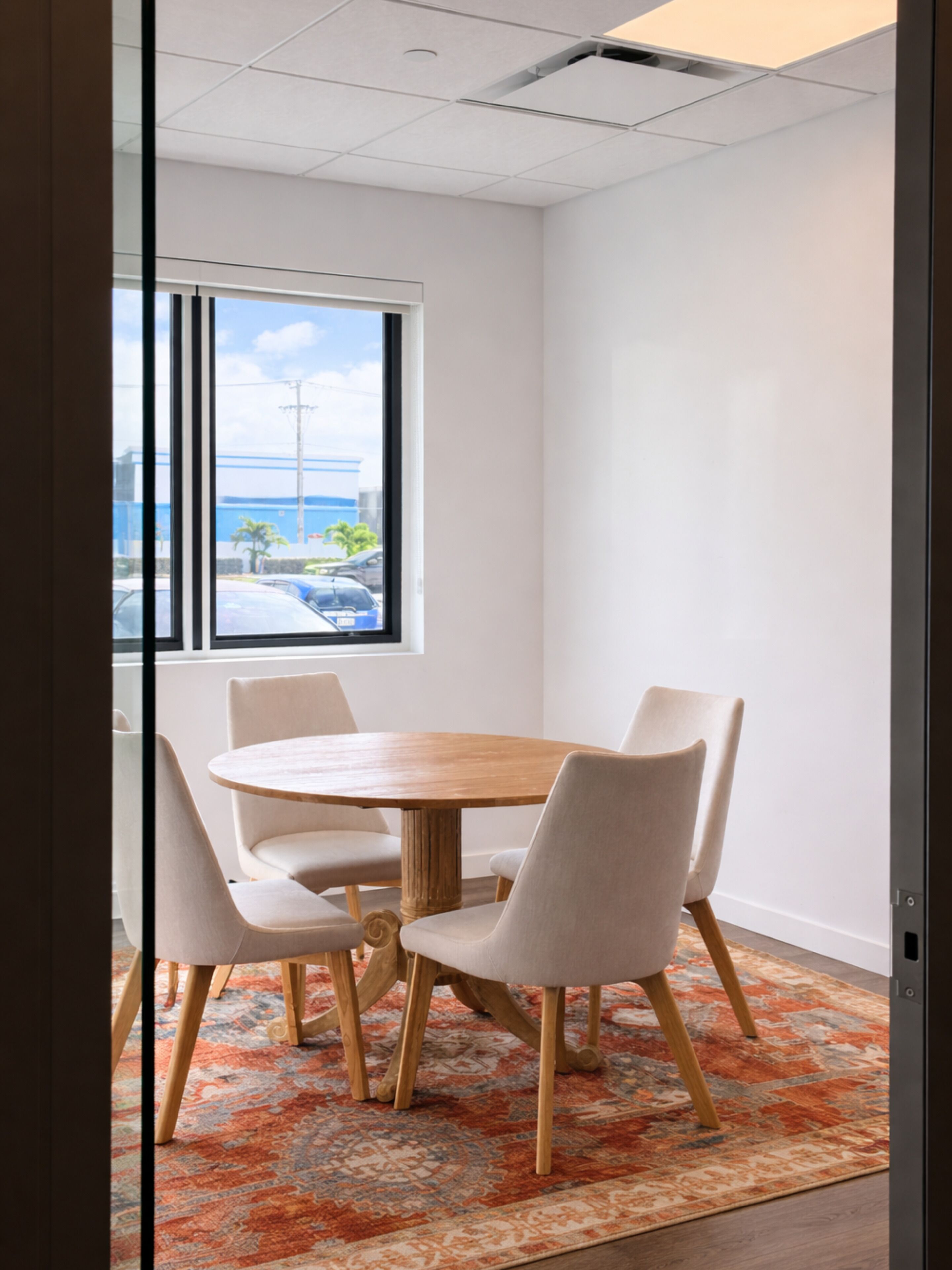 Ein Konferenzraum mit rundem Tisch, vier Stühlen, einem gemusterten Teppich und Blick auf ein Gebäude und Palmen durch ein Fenster.