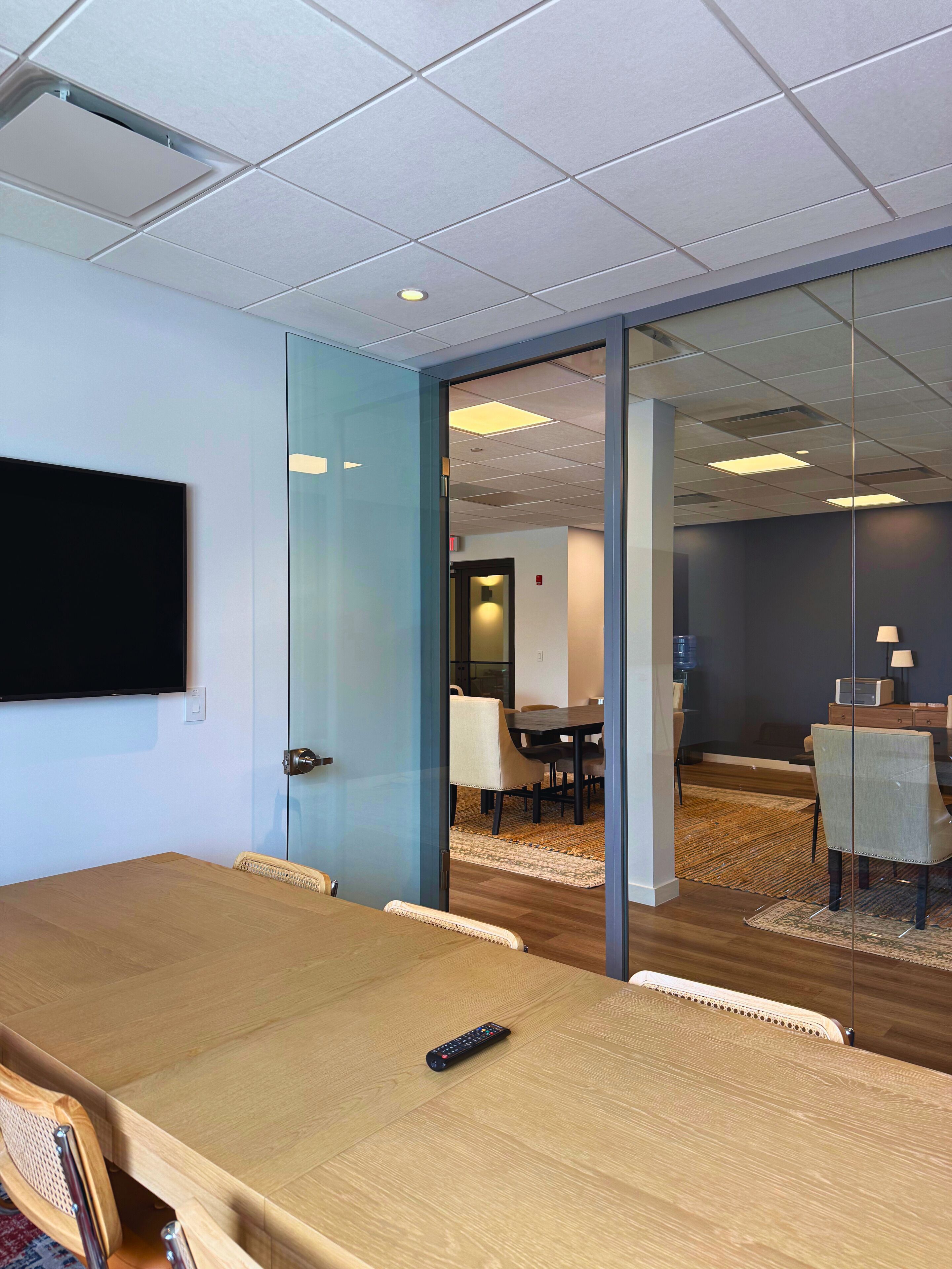 Un salón de conferencias con una mesa grande, sillas, un televisor montado en la pared y una puerta de cristal que da a otra sala.