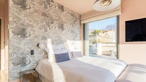 Ein modernes Schlafzimmer mit einer Wand im Steinoptik, einem großen Bett mit weißer Bettwäsche und Blick ins Freie durch ein großes Fenster.