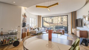 Ein modernes Wohnzimmer mit einem runden Esstisch, grünen Stühlen, einer Couch und einem großen Fenster mit Blick auf ein historisches Gebäude.