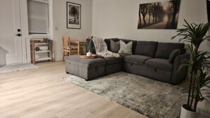 Een moderne woonkamer met een grijze hoekbank, een houten eettafel met stoelen, een klein bijzettafeltje en een potplant.