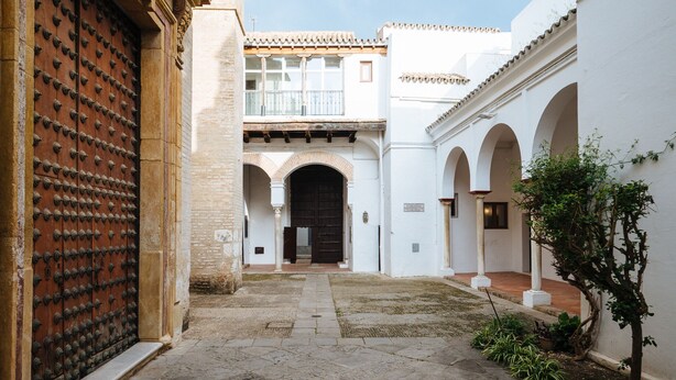 Un patio con un gran puerta de madera, pasillos en arco y un pequeño árbol.