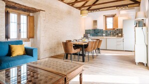 Amplia sala de estar con mesa de comedor, sofá azul y mesa de centro de vidrio. Se observa una cocina al fondo, con salpicadero de azulejos y armarios de madera.