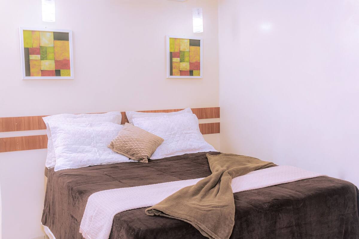 A neatly made bed with a brown cover, white pillows, and a beige throw. There are two abstract paintings on the wall above the bed.