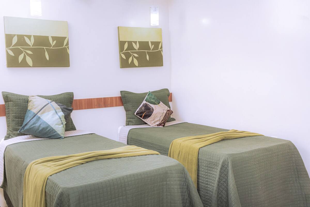 A hotel room with two beds, each with green bedding and a yellow throw blanket. There are two wall-mounted artworks depicting branches.