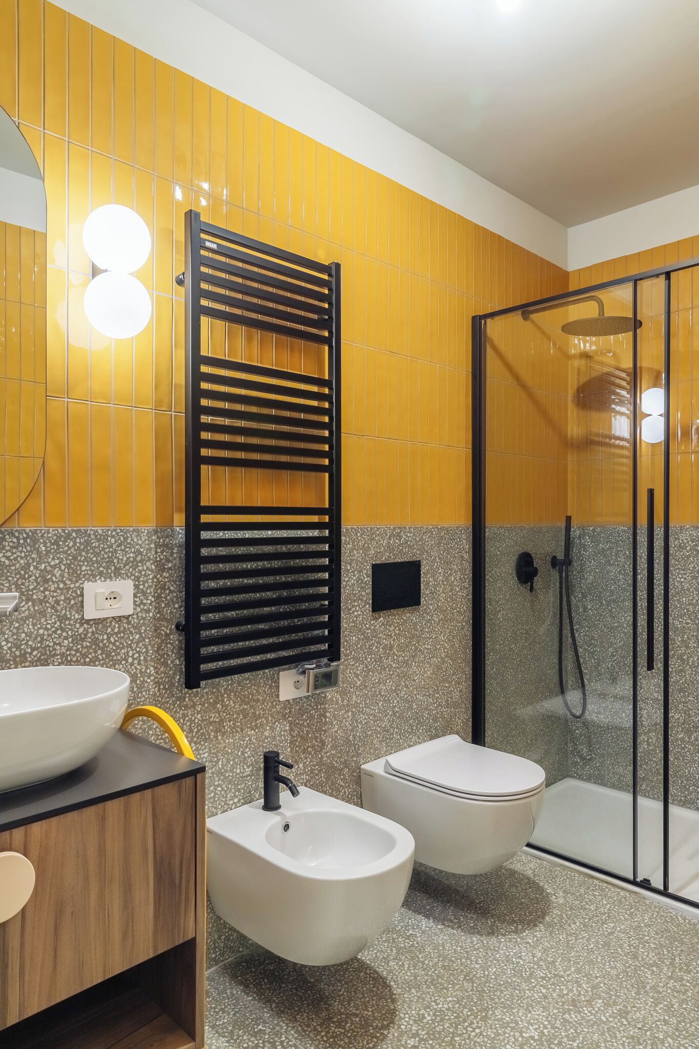 Ein modernes Badezimmer mit gelben Fliesen, einem schwarzen Handtuchhalter, einem weißen Waschbecken, einem Bidet und einer Duschkabine aus Glas.