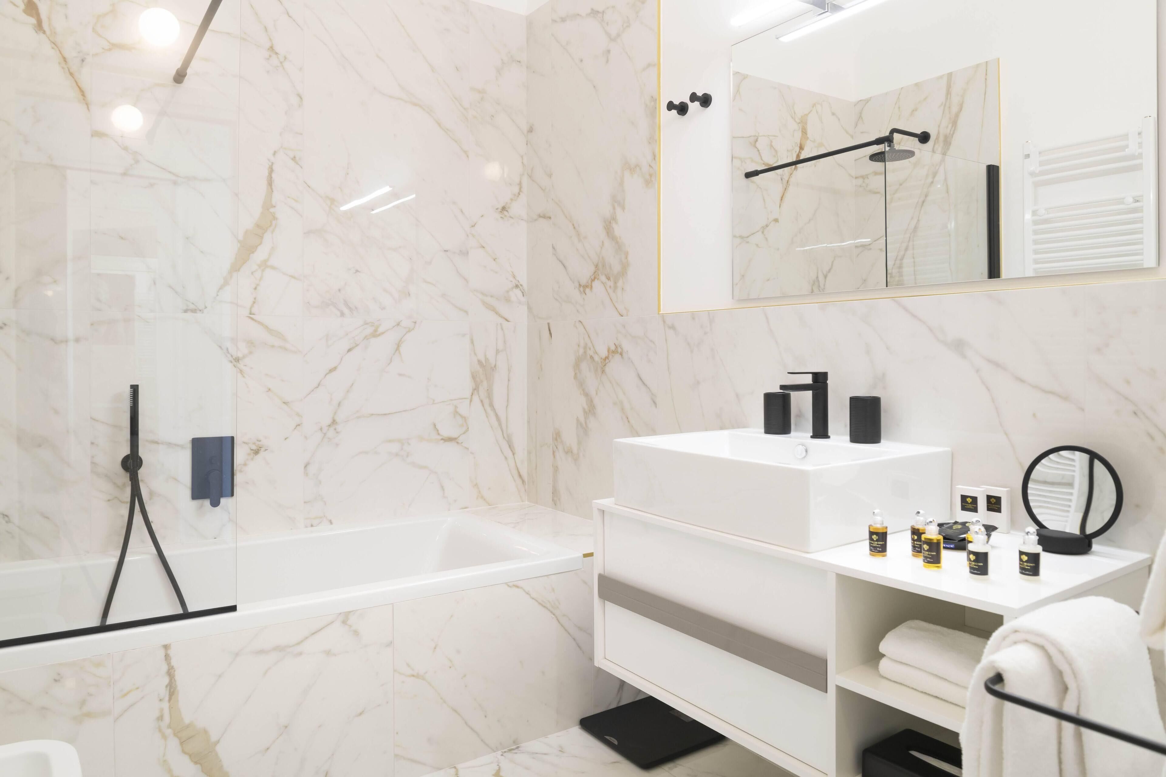 A modern bathroom with a marble wall, a bathtub, a vanity with toiletries, and a mirror.