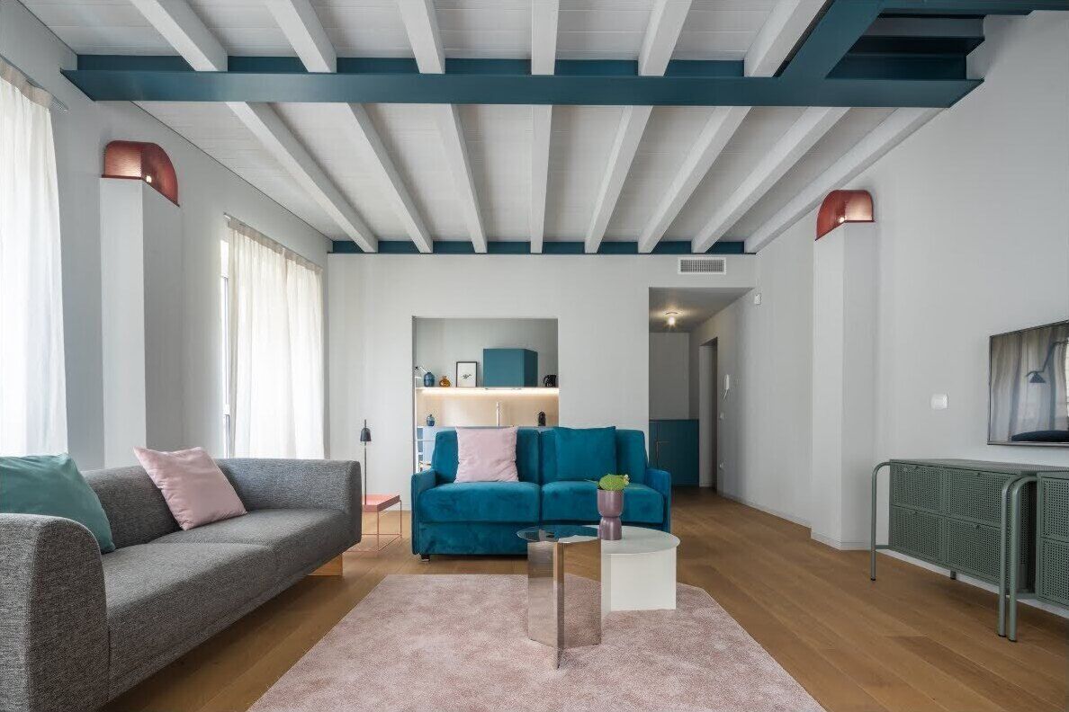 A modern living room with a grey sofa, a teal sofa, and a coffee table. There is a kitchen area in the background with blue cabinets and a white countertop.