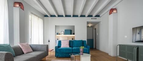 A modern living room with a grey sofa, a teal sofa, and a coffee table. There is a kitchen area in the background with blue cabinets and a white countertop.