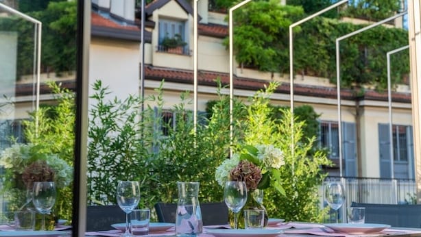 Un espace de restauration en plein air avec une table dressée pour deux personnes, entouré de verdure et offrant une vue sur les bâtiments.