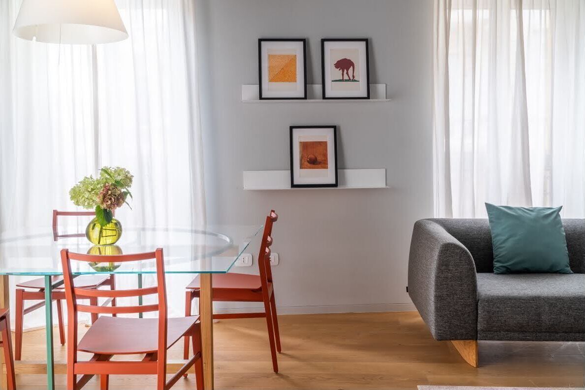 A modern living room with a glass dining table, red chairs, a grey sofa with a teal pillow, and framed artwork on the wall.