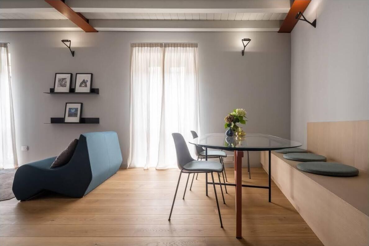 A modern living room with a glass dining table, a blue sofa, and a small seating area with cushions.