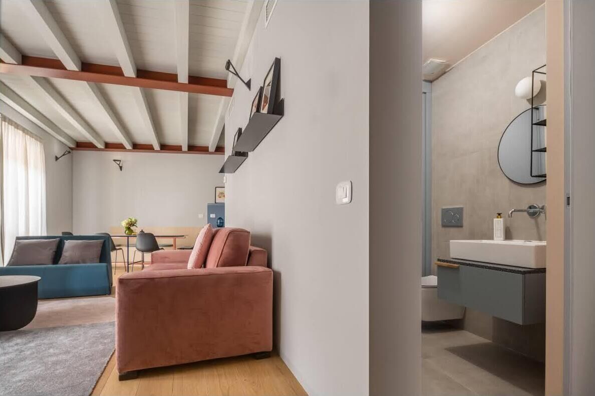 Ein modernes Wohnzimmer mit einem rosafarbenen Sofa, einem blauen Sofa und einem runden Couchtisch. Ein kleiner Tisch mit Vase und Bilderrahmen steht ebenfalls dort. Das Zimmer hat eine Holzbalkendecke und eine Leuchte.