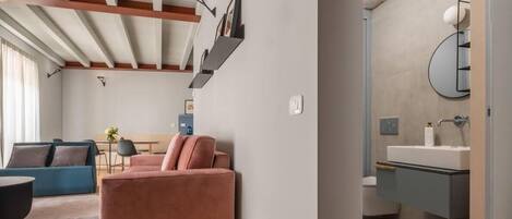 Ein modernes Wohnzimmer mit einem rosafarbenen Sofa, einem blauen Sofa und einem runden Couchtisch. Ein kleiner Tisch mit Vase und Bilderrahmen steht ebenfalls dort. Das Zimmer hat eine Holzbalkendecke und eine Leuchte.