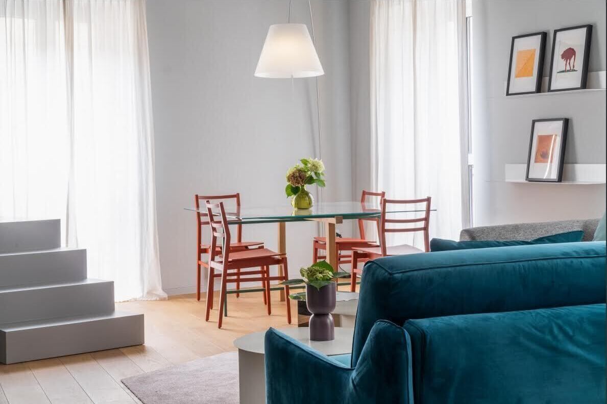 Een moderne woonkamer met een blauwgroene bank, een glazen eettafel met stoelen, een witte lamp en een trap.
