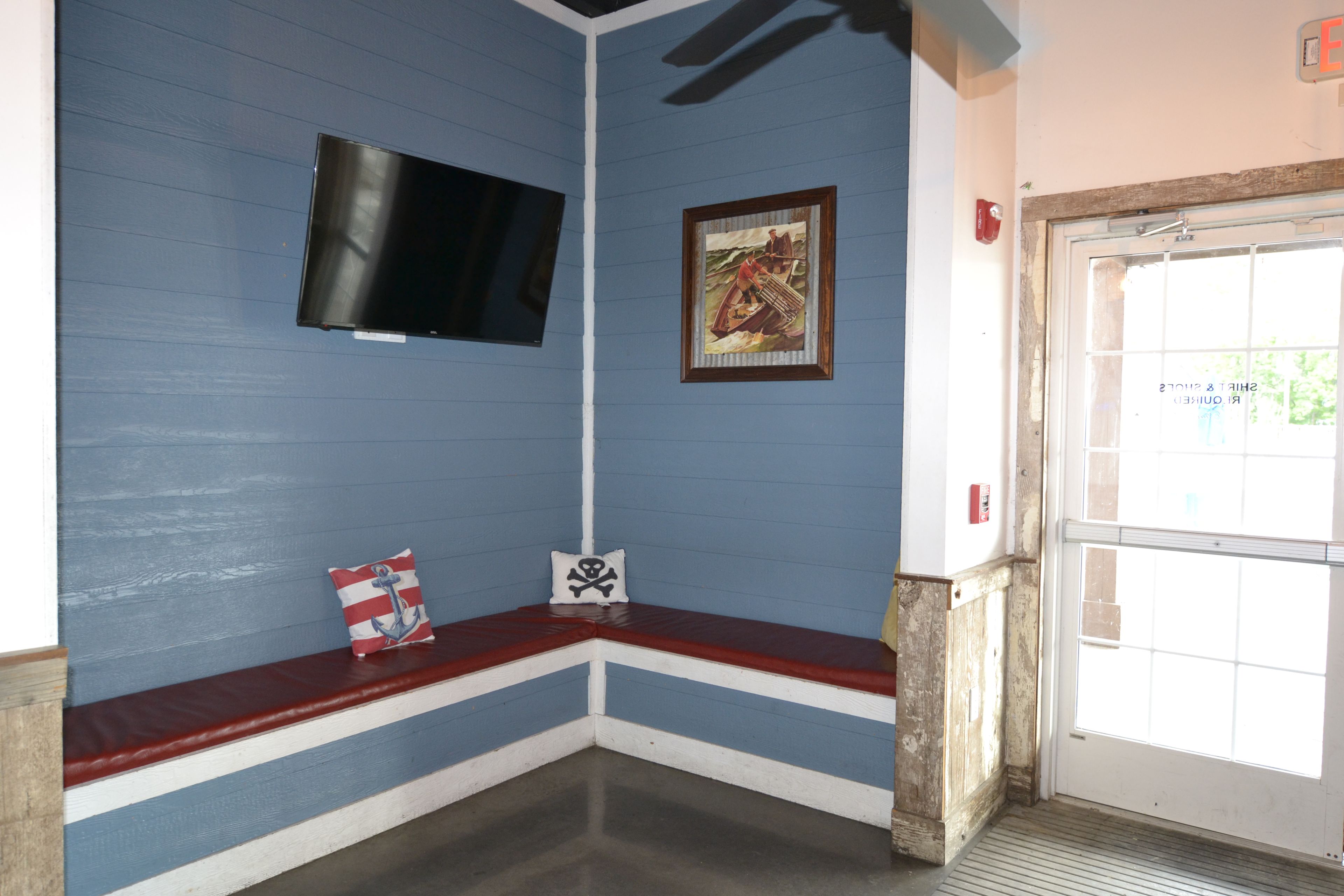 A room with a blue wall, a mounted television, a framed picture, a bench with pillows, and a door with a window.
