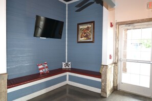 Une pièce avec un mur bleu, un téléviseur fixé au mur, un tableau encadré, un banc avec des coussins et une porte avec une fenêtre.
