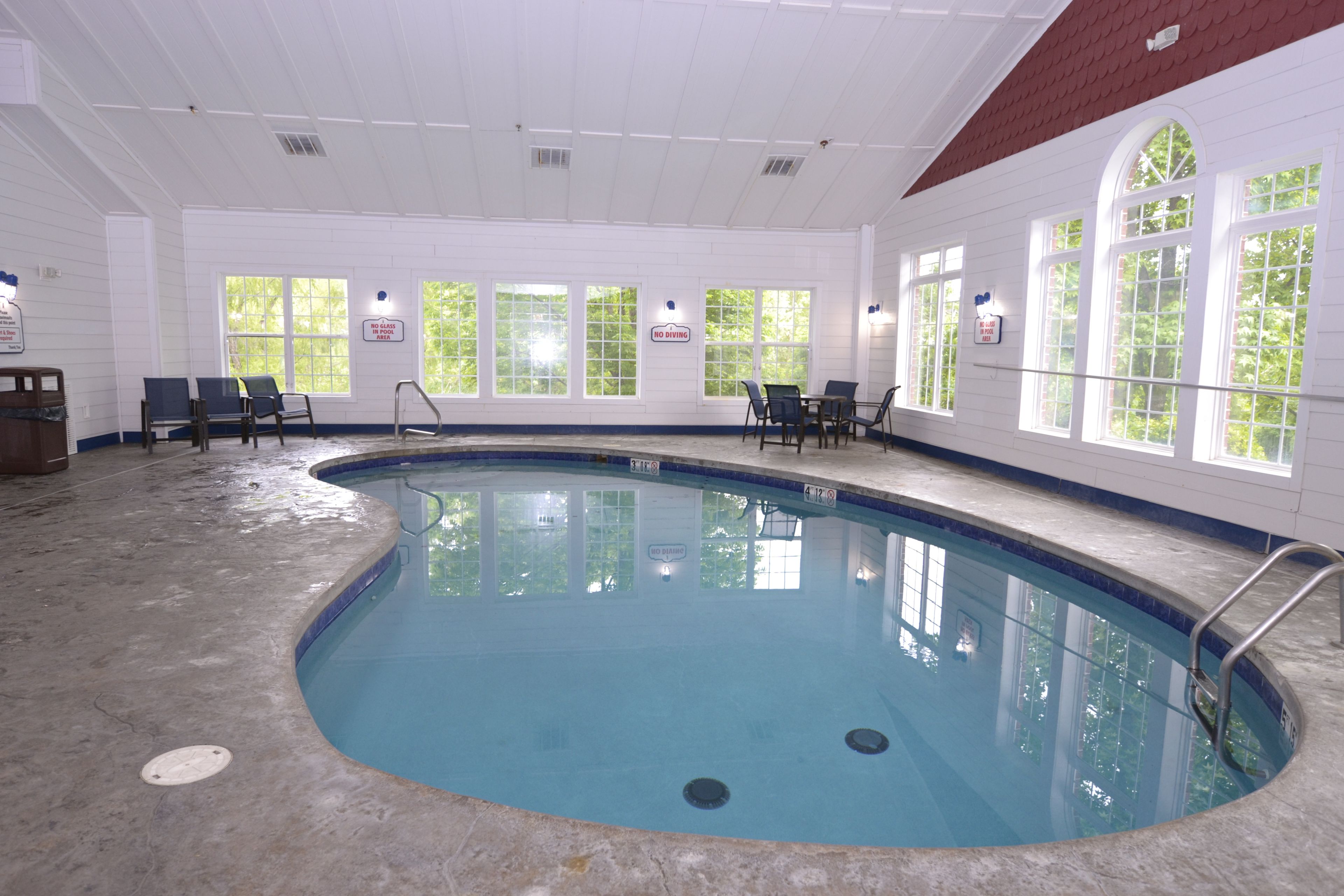 Una piscina interior con ventanas amplias, una escalera de piscina y algunas sillas alrededor.