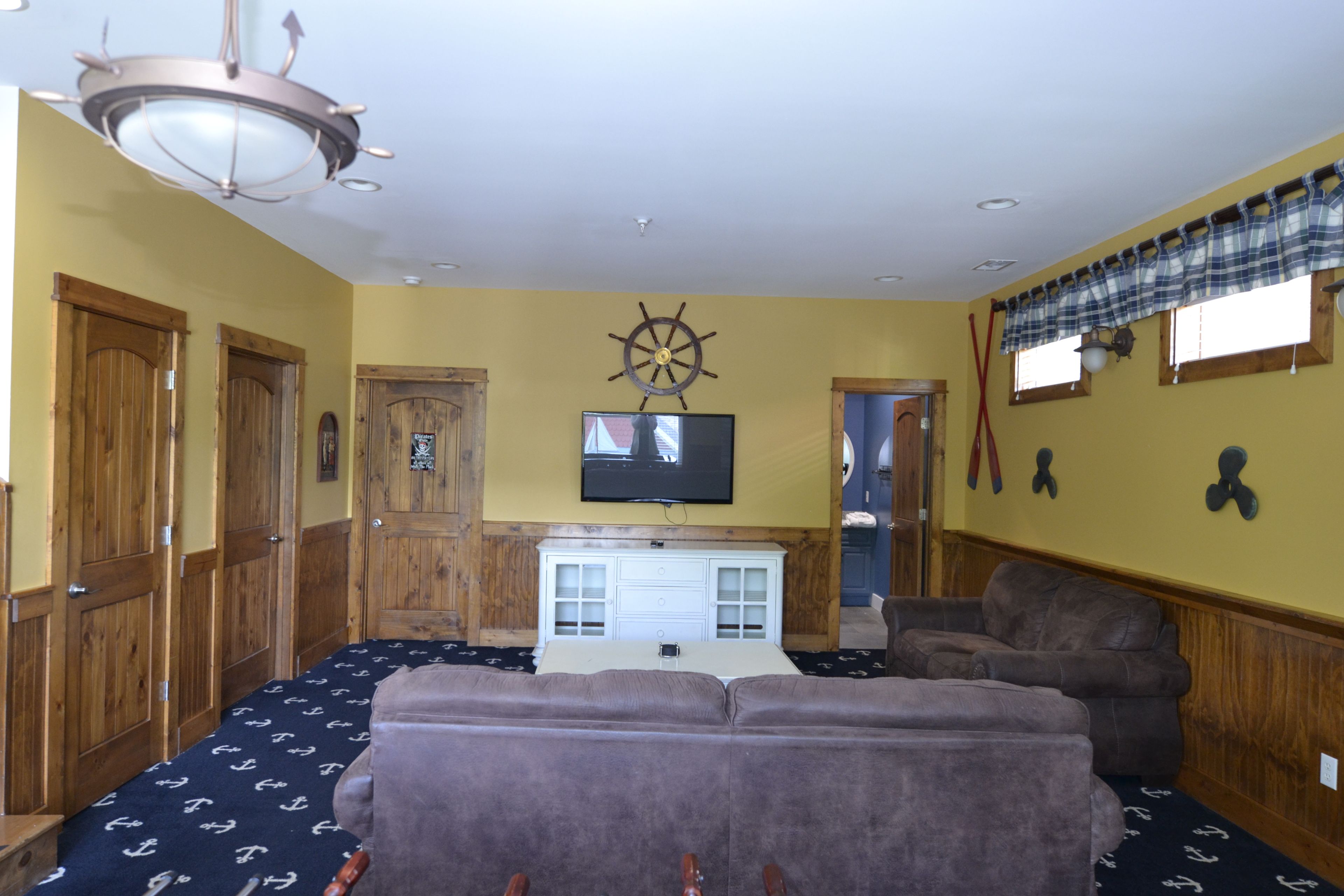 A room with wooden doors, a television mounted on the wall, a ceiling fan, and a window with a plaid valance.
