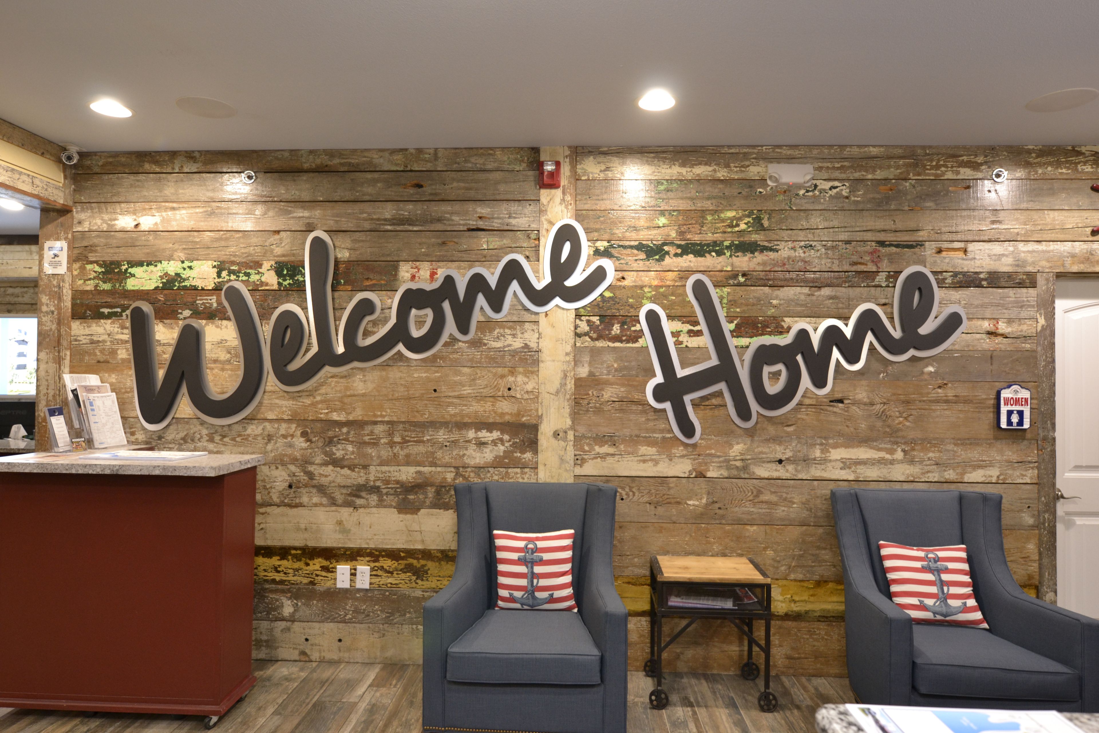 A reception area with a wooden wall and a 'Welcome Home' sign. There are two armchairs with striped pillows and a small table.