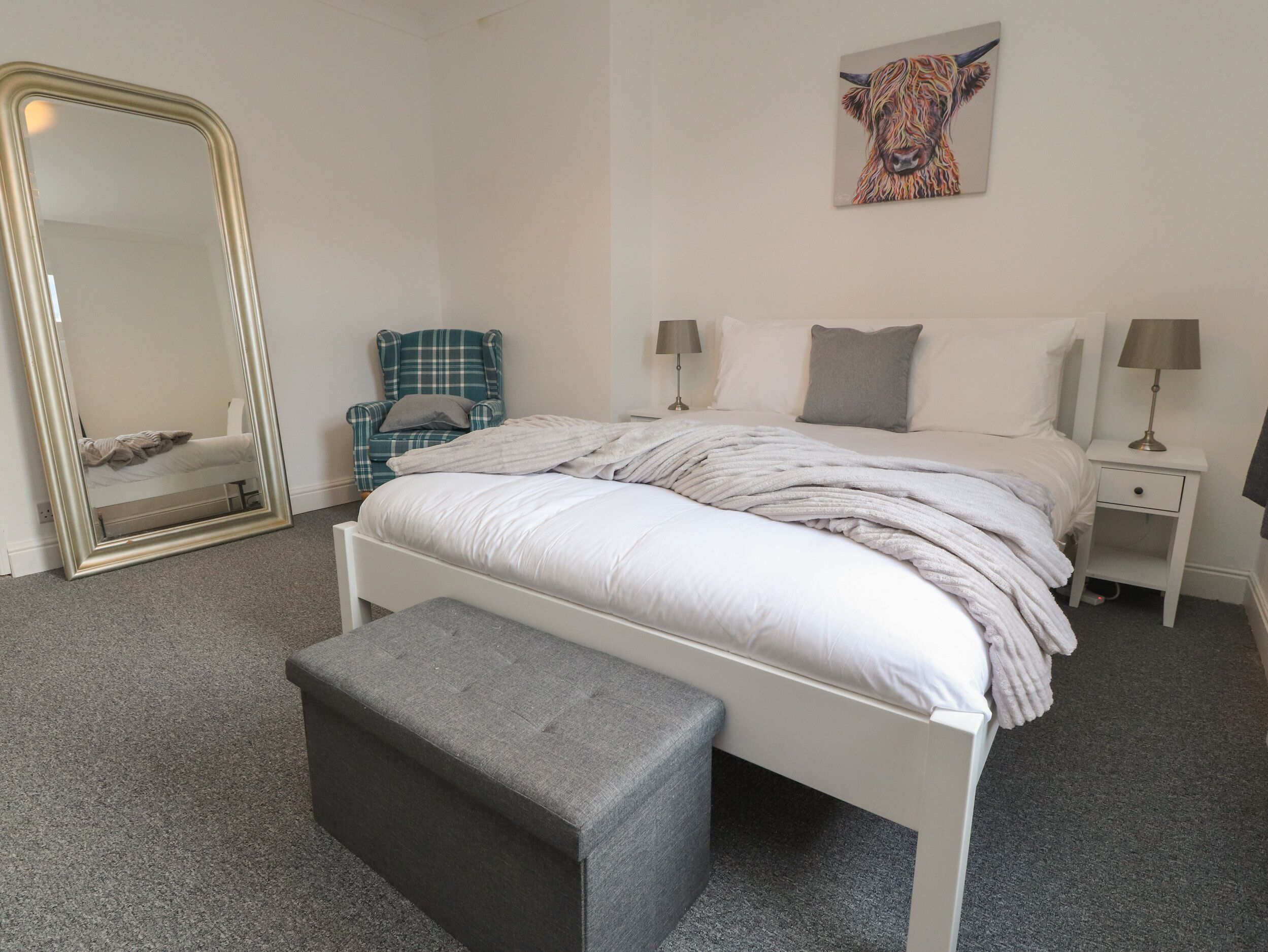 Een slaapkamer met een grote spiegel, een bed met witte lakens, een bankje, een stoel en een schilderij aan de muur.