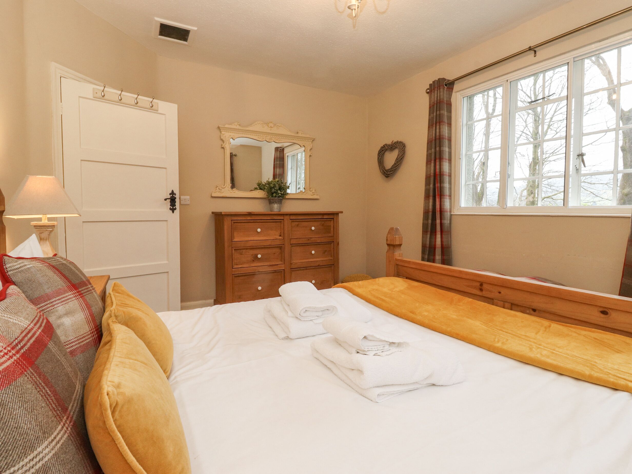 Een keurig opgemaakt bed met witte lakens en handdoeken, een houten dressoir met meerdere lades, een spiegel boven het dressoir, een lamp op een nachtkastje, en een raam met gordijnen.