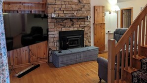 A living room with a stone fireplace, wooden flooring, a television, and a staircase. - The Lakeshore House (Niverville)