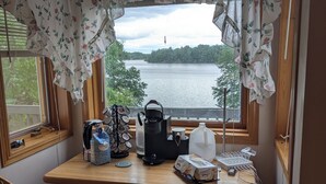 A corner room with a wooden desk, a chair, a coffee maker, a jug, and a view of a lake through floral curtains. - The Lakeshore House (Niverville)