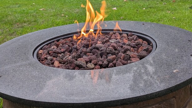 A circular stone fire pit with flames, surrounded by a grassy area and a house in the background. - The Lakeshore House (Niverville)