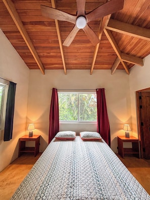 A bedroom with a wooden ceiling, a bed with patterned bedding, two bedside tables with lamps, and a window with curtains.