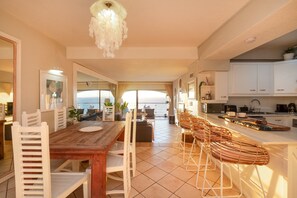Una cocina amplia con una mesa de comedor de madera, sillas blancas y vistas al mar a través de ventanas grandes.
