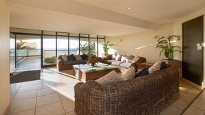 A spacious living area with wicker furniture, a glass coffee table, and a view of the sea through large windows.