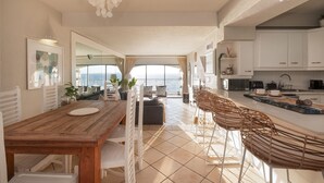 A modern kitchen with white cabinets, a wooden dining table, and a large window overlooking the sea.