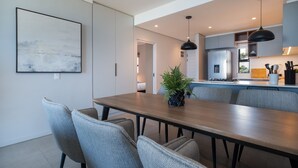 A modern dining area with a wooden table and grey upholstered chairs, a potted plant, and a kitchen in the background. - Umhlanga Ridge | Pool | Sauna | Aircon |Gym (Cape Town)