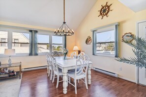 Uma área de refeições com mesa e cadeiras brancas, um candelabro e janelas amplas com vista para edifícios.