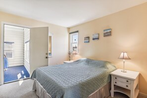 Quarto com cama, mesa de cabeceira, abajur, janela e porta para outro espaço.