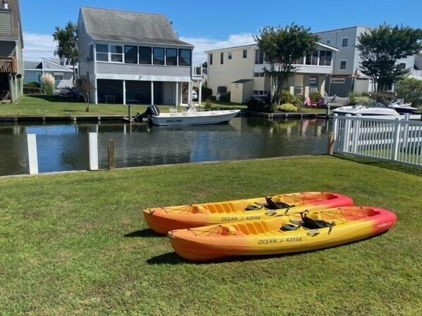 Two kayaks on a grassy area near a body of water with boats docked in the background.
