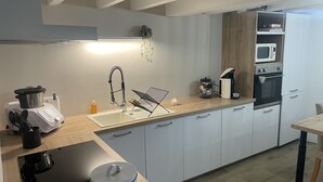 Een moderne keuken met een inbouwfornuis, magnetron en oven. Er is een spoelbak met een moderne kraan, een aanrecht met een waterkoker en een kaars, en een kleine plant aan de muur.