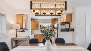 Comedor con mesa de madera, cuatro sillas, un florero blanco con plantas y una lámpara colgante.