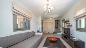 Ein modernes Wohnzimmer mit grauem Sofa, Couchtisch und Fernseher auf einem schwarzen Schrank. Eine Treppe führt in einen anderen Raum und ein Fenster mit Jalousien ist vorhanden.