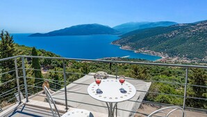 Eine Terrasse mit Tisch und zwei Gläsern Rotwein mit Blick auf eine Küstenlandschaft mit Bergen und einem klaren, blauen Meer.