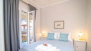 Ein ordentlich bezogenes Bett mit floralem Überwurf, zwei Kissen mit Zickzackmuster, ein gerahmtes Bild an der Wand und ein Balkon mit Blick auf Gebäude.