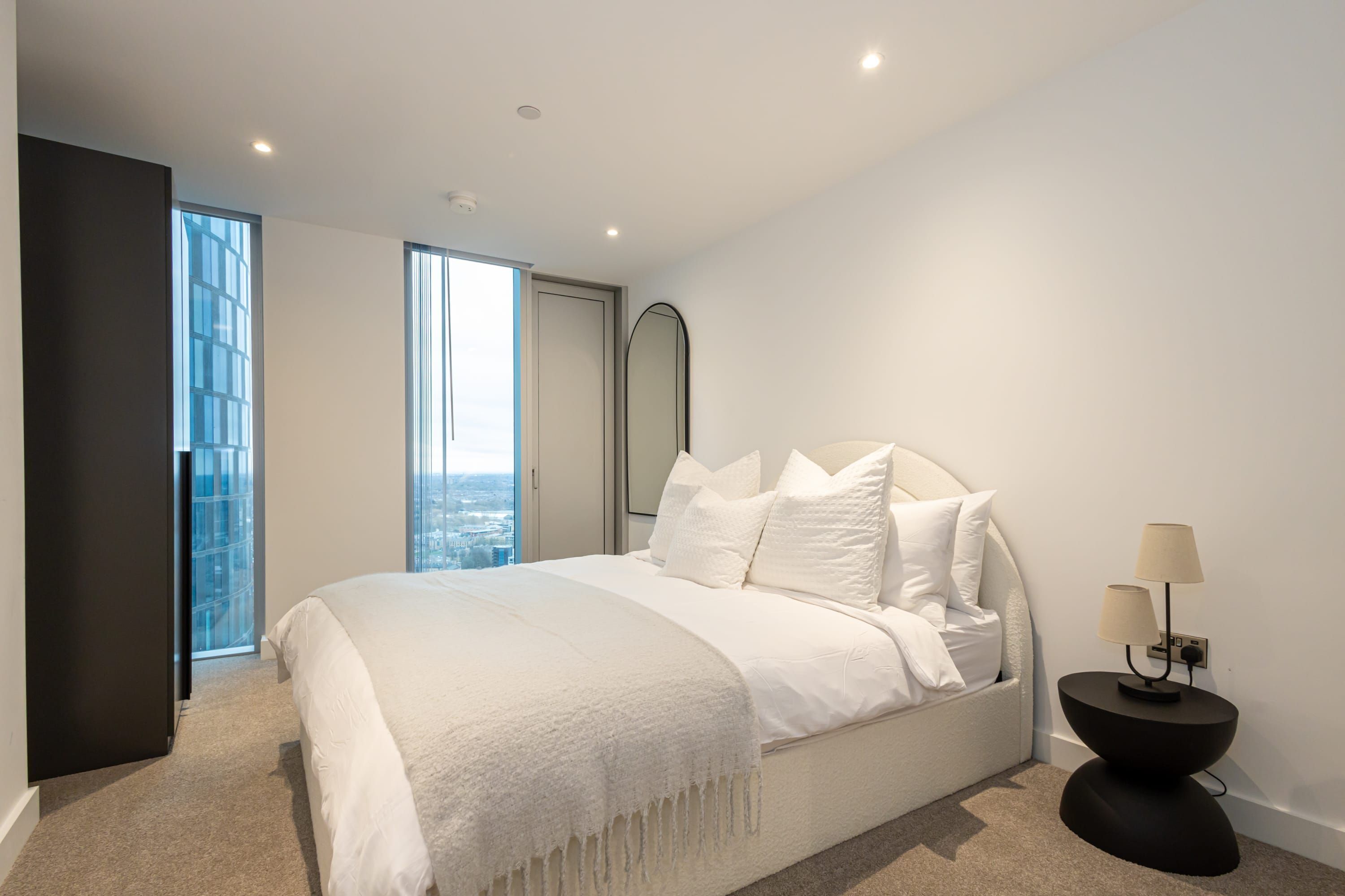 A modern bedroom with a large bed, a nightstand, and a view through a large window.
