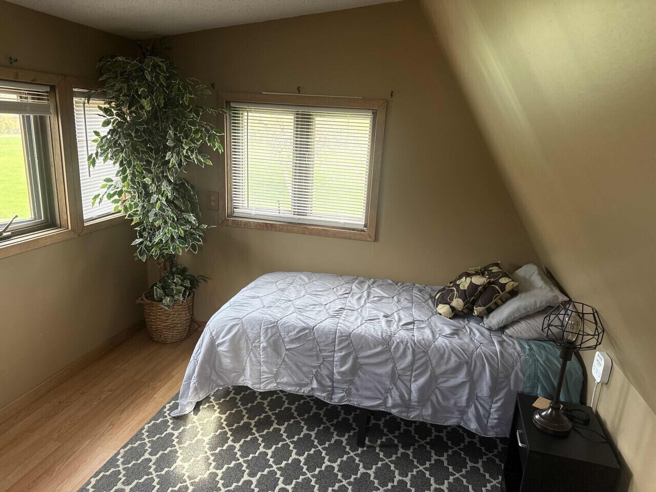 Un dormitorio con cama, planta en maceta, ventana y mesita de noche.