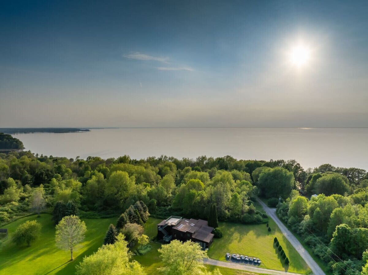 An aerial view of a property with a large body of water in the background, surrounded by dense greenery and a single building with a sloped roof.