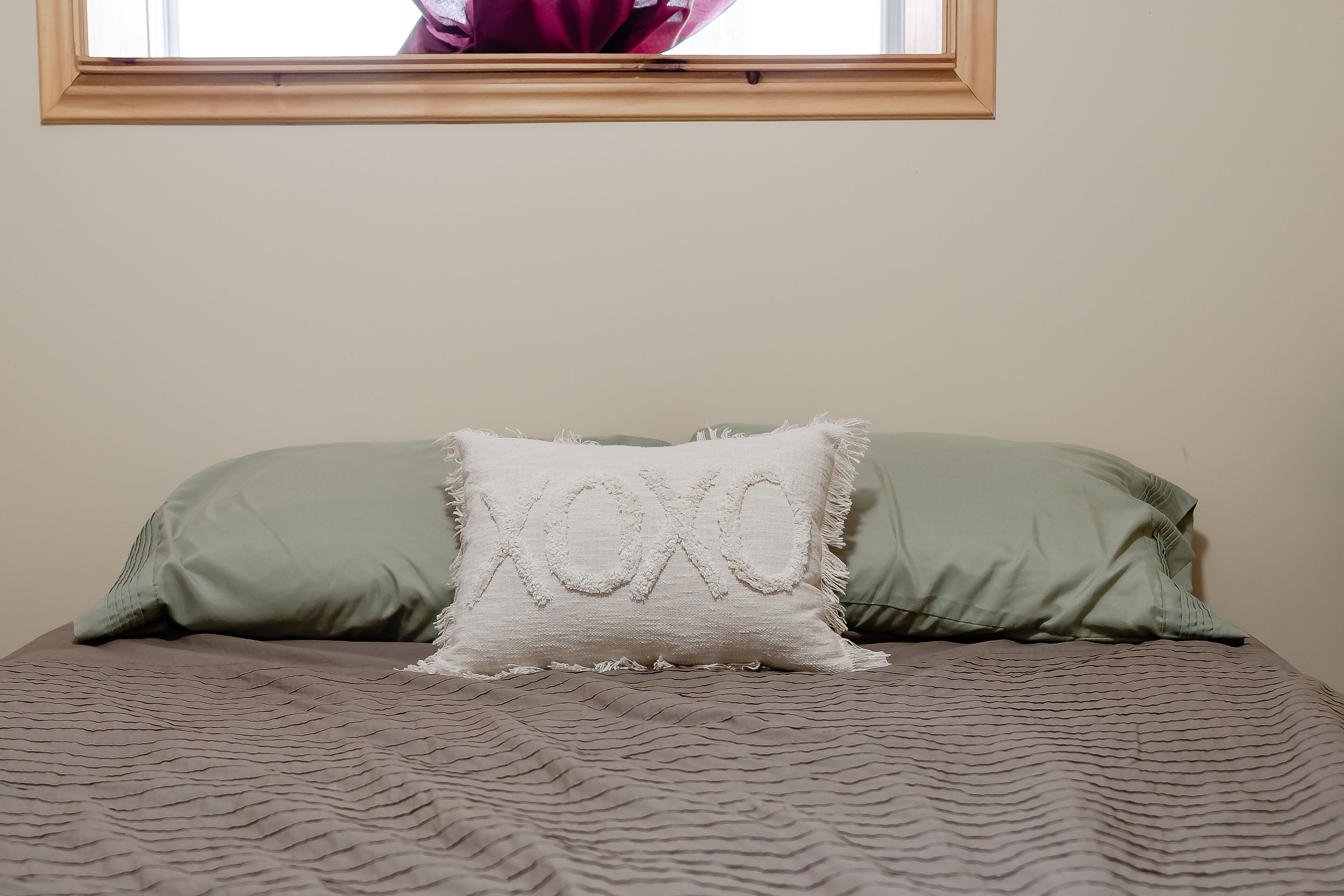 Un lit recouvert d’une couverture marron à motifs, avec un oreiller vert et un oreiller blanc orné d’un motif « XOXO » brodé.