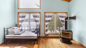 A room with a bed, a window with curtains, a wooden floor, and a wood-burning stove.