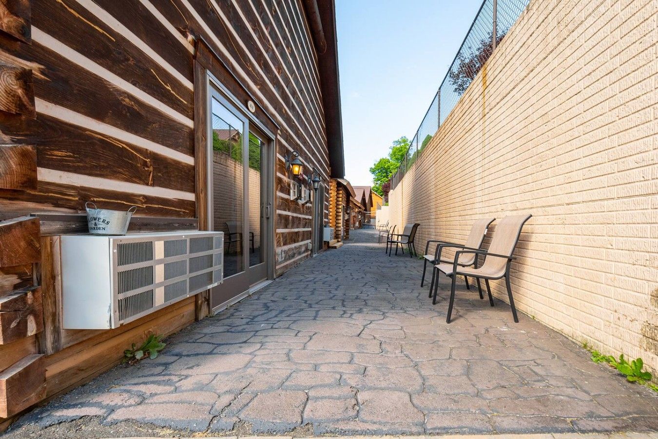 A narrow outdoor corridor with a wooden building on one side and a brick wall on the other. There are outdoor seating chairs and a small table with a metal container.