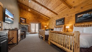 Une pièce aménagée dans le style d’un chalet en rondins, avec une cheminée, un lit doté d’une tête de lit en bois, un bureau avec un ordinateur et un téléviseur fixé au mur.
