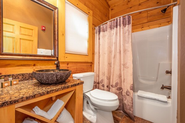 A bathroom with a wooden interior, a granite countertop, a white toilet, and a shower with a floral curtain. - Mountainside Cabin 40L-Dog Friendly (McHenry)